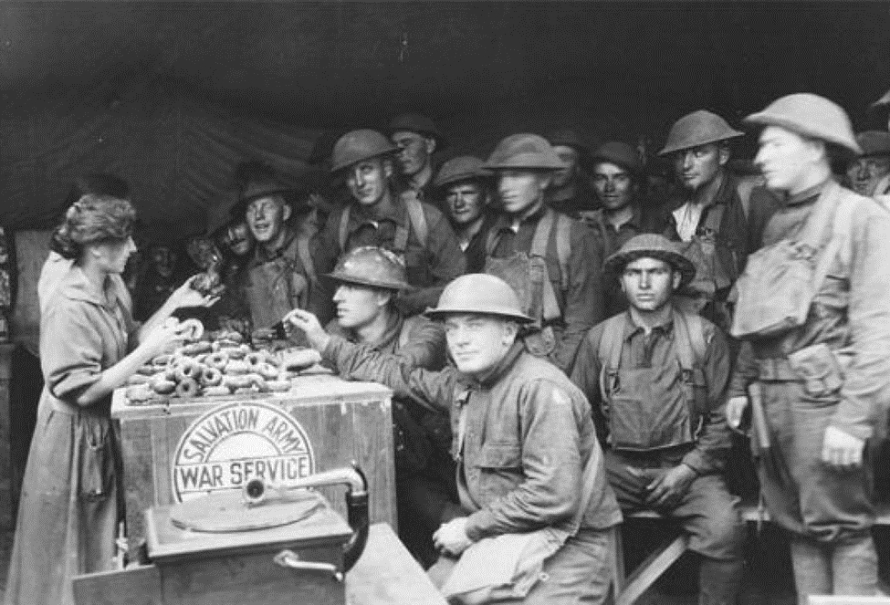 Foto antigua de la primera guerra mundial, en donde una chica adolescente y sonriente, sostiene un contenedor con muchas donas.