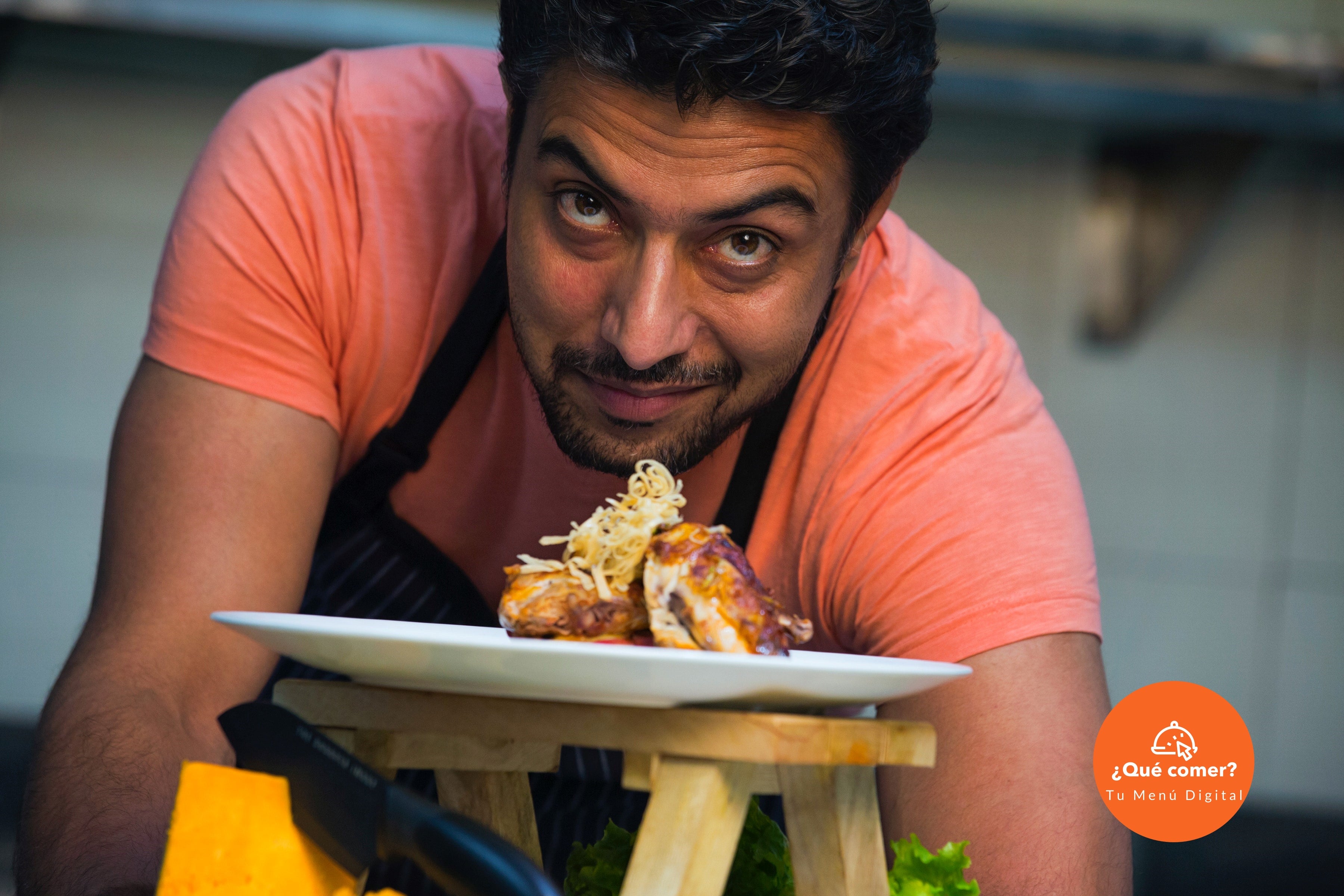 Hombre joven, atractivo, de tez morena y ojos grandes; inclinado frente a ti, mirándote fijamente y ofreciéndote un plato con un delicioso pollo dorado.