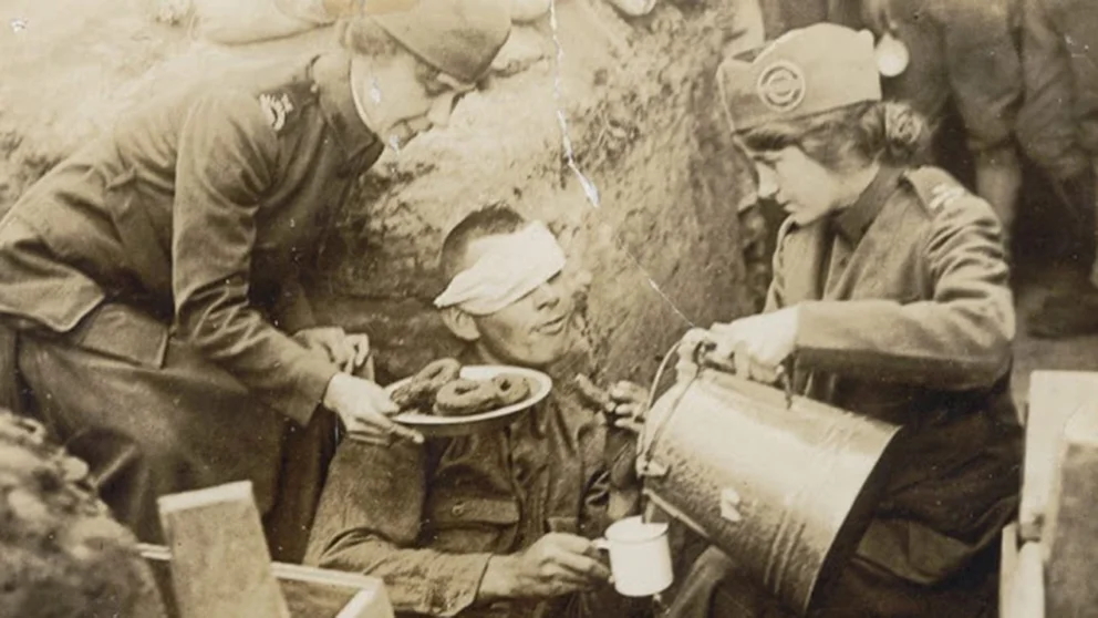 Foto antigua de la primera guerra mundial, en donde dos mujeres se encuentran en trinchera ofreciendo café y donas a un soldado que se encuentra herido con la cabeza vendada y la venda cubre uno de sus ojos.